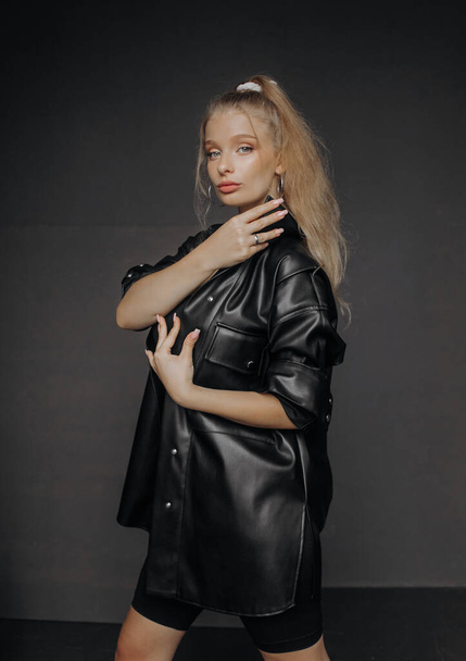 Amazing girl with blond hair dressed in a stylish top, shorts and leather shirt posing on a dark background. - Foto, imagen