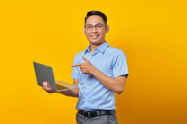 Портрет веселого красивого азиата в очках, указывающего пальцем на ноутбук, показывающего что-то в сети изолированное на желтом фоне. бизнесмен и предприниматель концепция - Фото, изображение