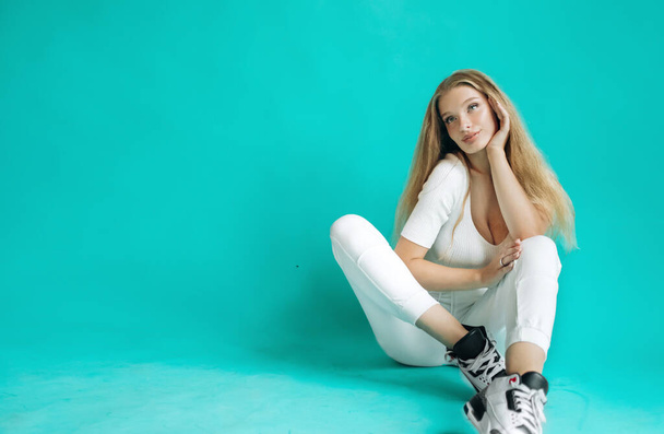 Luxus-Blondine in weißer Sportbekleidung posiert im Studio auf türkisfarbenem Hintergrund. - Foto, Bild