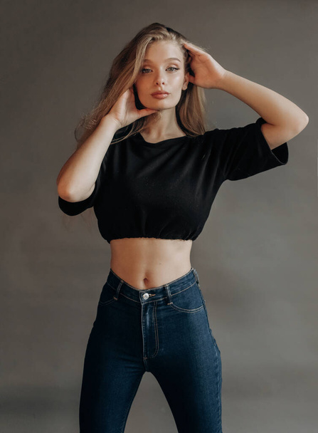 Сексуальная блондинка в черном топе и джинсах позирует в студии на сером фоне - Фото, изображение