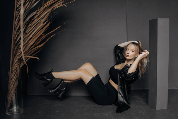 Stylish blonde sitting on the floor raises her legs together, posing in the studio on a gray background. - Φωτογραφία, εικόνα