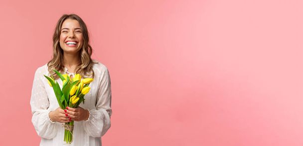 Urlaub, Schönheit und Frühlingskonzept. Porträt von glücklich aufgeregt charmante blonde Mädchen erhalten Blumen, kaufen gelbe Tulpen selbst, lächeln und lachen freudig, stehen rosa Hintergrund - Foto, Bild