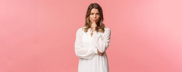 Beauty, fashion and women concept. Portrait of skeptical and judgemental serious-looking blond female in white spring dress, look disbelief, grimacing and frowning, pink background - Foto, immagini