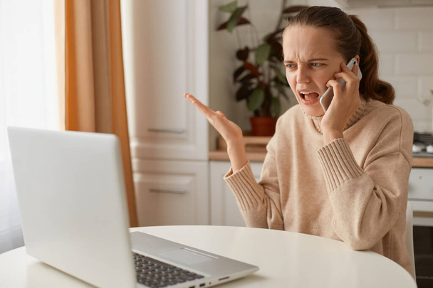 Indoor shot of confused woman with dark hair wearing beige sweater sitting in kitchen and working on laptop, holding cell phone and having unpleasant conversation. - Photo, image
