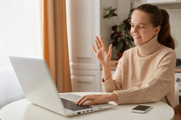 Indoor shot of adorable smile female woman with dark hair wearing beige sweater sitting in kitchen and working on laptop, having online conversation, video call. - Foto, Bild