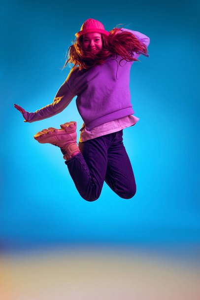 Portrait of young stylish girl posing in a jump isolated over blue studio background in pink neon light - Фото, зображення