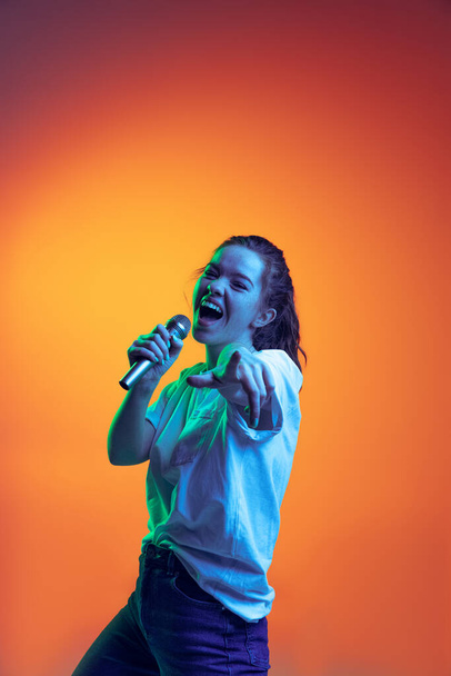 Portrait of young girl emotively singing in microphone, posing isolated over gradient red background in neon light. Music lifestyle - Фото, изображение