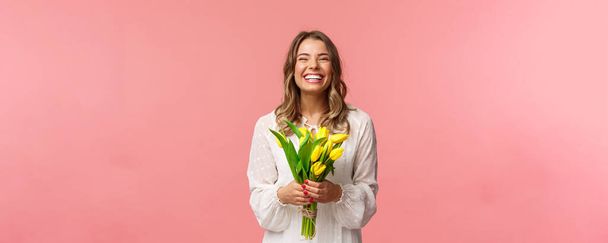 Vacaciones, belleza y concepto de primavera. Retrato de niña rubia encantadora emocionada feliz recibir flores, la compra de tulipanes amarillos a sí misma, sonriendo y riendo alegremente, de pie fondo rosa
 - Foto, Imagen
