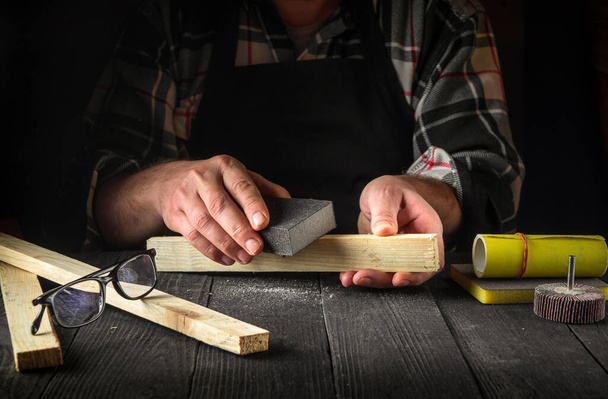A carpenter polishes a wooden plank with an abrasive tool. Hands of the builder close-up during work. Renovation or construction idea - Photo, Image