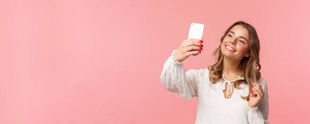 Крупним планом портрет красивої і стильної блондинки, яка весною приймає селфі в парку, збирає любителів і послідовників, робить блогерський контент, фотографує на смартфоні, посміхається пухнастий, рожевий фон
 - Фото, зображення