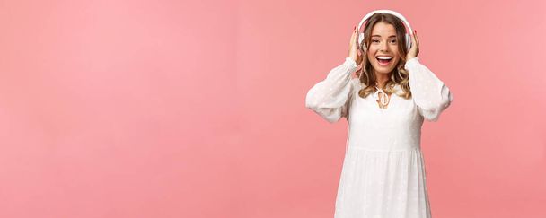 興奮し、幸せな良い白い柔らかいドレスの少女の肖像画は、ヘッドフォンを着用し、笑顔カメラを見て、良い音質で魅了し、ピンクの背景に驚いて - 写真・画像