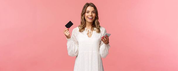Portret beztroskiej młodej ładnej blondynki korzystającej z telefonu komórkowego, aby zapłacić za zakup online, trzymając kartę kredytową i uśmiechnięty aparat fotograficzny, zakupy internet, pobierz aplikację na smartfona - Zdjęcie, obraz