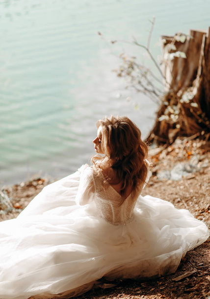 Η νεαρή νύφη κάθεται κοντά στη λίμνη στον ήλιο, χαϊδεύοντας το πρόσωπό της. - Φωτογραφία, εικόνα