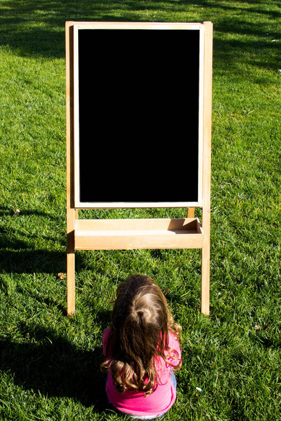 adorable niña sentada en el jardín mirando una pizarra donde se puede insertar texto o imágenes - Foto, imagen