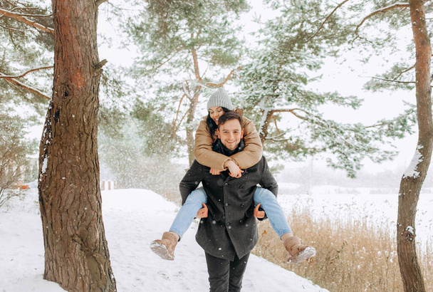 Chlápek jezdí na zádech na své přítelkyni, zatímco se prochází ve sněženém lese. Zimní dovolená, odpočinek. - Fotografie, Obrázek