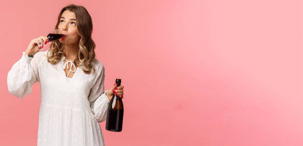Holidays, spring and party concept. Portrait of young elegant happy blond woman, wear white trendy dress, drinking wine from glass looking up pleased, tasting good drink, hold bottle - Photo, Image