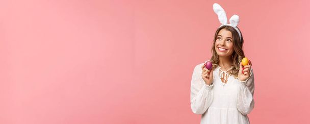 Holidays, spring and party concept. Portrait of dreamy, tender young blond girl feeling happy, celebrating religious day, holy Easter, wear rabbit ears and white dress, hold painted eggs, look away - Фото, зображення