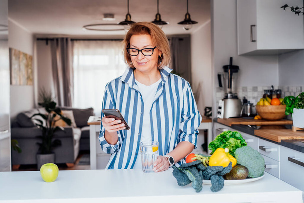 Healthy meal diet planning. Woman making notes, type her menu in phone app sitting above the table with fresh vegetables on her kitchen. Vegetarian, plant-based foods. New healthy lifestyle habits. - 写真・画像