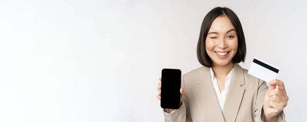 Усміхнена корпоративна жінка в костюмі, показує екран мобільного телефону і додаток на мобільному телефоні, екран смартфона, що стоїть на білому тлі
 - Фото, зображення