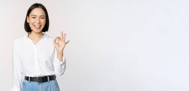 Very good. Smiling asian woman showing okay with satisfied face expression, praise and compliment great job, pleased by smth, standing over white background - Photo, Image