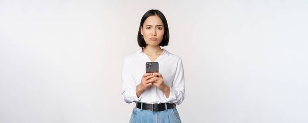 Asiatin mit Smartphone und zweifelndem Blick, enttäuscht von Handy-App, vor weißem Hintergrund - Foto, Bild