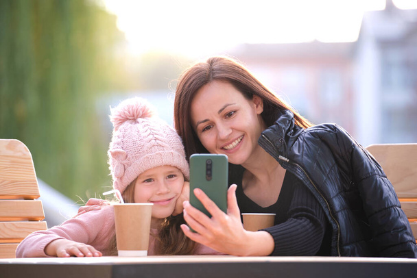 Young pretty woman taking picture with phone selfie camera with her child daughter sitting at street cafe with hot drinks on sunny autumn day. Happiness in family relations concept - 写真・画像