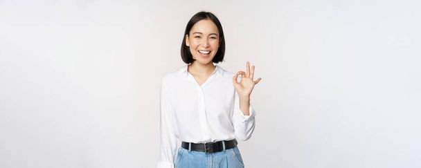 Heel goed. glimlachende aziatische vrouw tonen oke met tevreden gezicht uitdrukking, lof en compliment groot werk, blij door smth, staande over witte achtergrond - Foto, afbeelding
