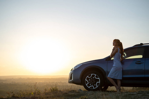 Conductrice heureuse en robe d'été bleue profitant d'une soirée chaude près de sa voiture. Concept de voyage et vacances - Photo, image