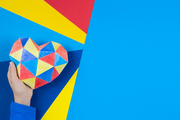 Mano del niño sosteniendo colorido corazón de papel sobre fondo de papel multicolor. Concepto del Día Mundial de la Conciencia del autismo - Foto, imagen