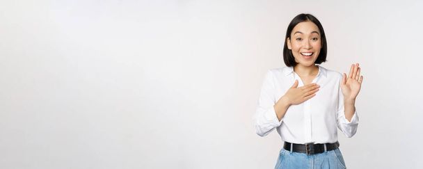 Εικόνα της χαριτωμένης νεαρής εργαζόμενης γραφείου, Ασιάτισσα φοιτήτρια που σηκώνει το χέρι και βάζει παλάμη στο στήθος, αυτοαποκαλείται, συστήνεται, δίνει υποσχέσεις, στέκεται πάνω από λευκό φόντο - Φωτογραφία, εικόνα