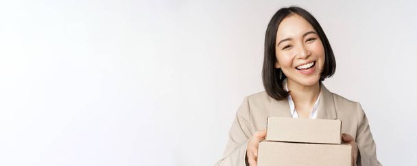 Image of asian saleswoman, business woman giving boxes with order, deliver to customer, standing in suit over white background - Photo, Image