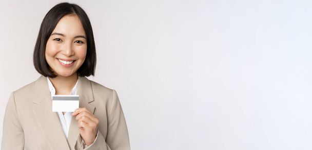 Commis de bureau souriant, femme d'affaires asiatique montrant carte de crédit, debout sur fond blanc en costume beige - Photo, image