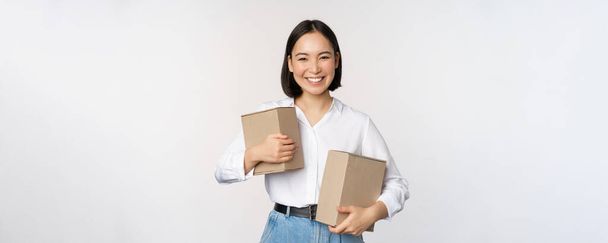 Концепція покупки та доставки. Молода щаслива азіатська жінка позує з коробками і посміхається, стоїть над білим тлом
 - Фото, зображення