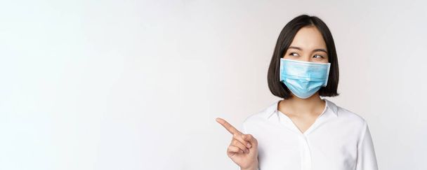 Ritratto di donna asiatica carina in maschera medica, protezione coronavirus, indicando il dito sinistro e guardando incuriosito lo spazio vuoto copia, sfondo bianco - Foto, immagini
