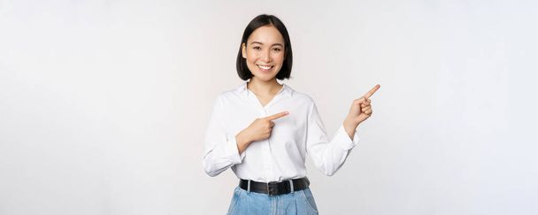 Портрет красивої азіатки, продавець вказує пальці праворуч і показує інформацію, банер або логотип, рекламу продажу, стоїть на білому тлі
 - Фото, зображення