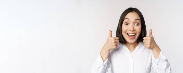 Enthusiastic asian girl showing thumbs up and smiling, pleased by something, recommending smth good, standing over white background - 写真・画像
