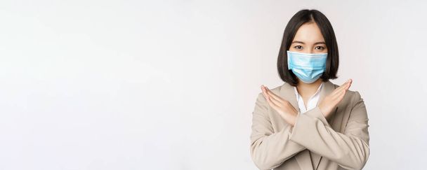 Coronavirus en werkplekconcept. Afbeelding van aziatische zakenvrouw, vrouw in medisch gezichtsmasker shows stop, kruis verbieden gebaar, staat over witte achtergrond - Foto, afbeelding