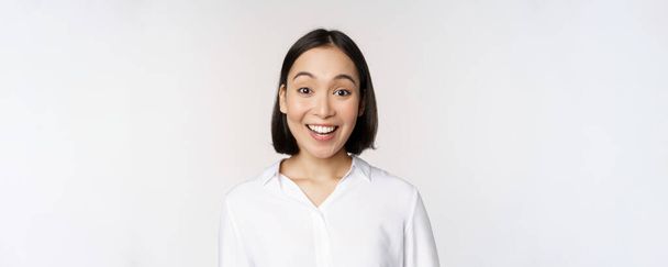 Close up portrait of young asian female model looking amazed at camera, smiling white teeth, standing against white background - Photo, Image