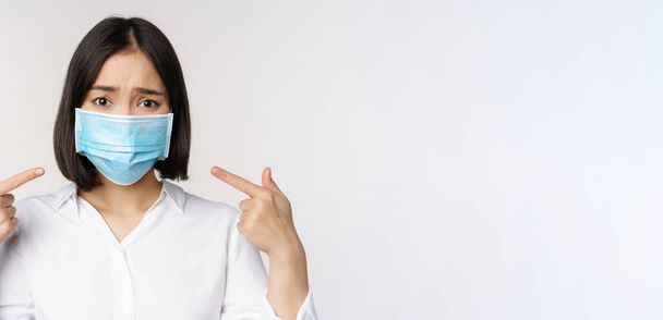 Close up of sad asian girl in medical face mask pointing at her head and looking upset, standing over white background - Photo, Image