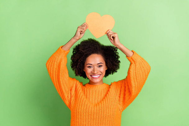 Photo of sweet young wavy hairdo lady hold heart wear orange sweater isolated on green color background - 写真・画像