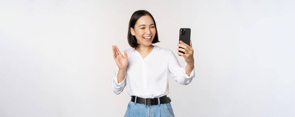 スマートフォンのフロントカメラで手を振ってスタイリッシュな現代アジアの女性のイメージ,ビデオチャット,携帯電話のアプリケーション上の人とチャット,白の背景 - 写真・画像