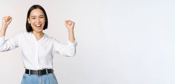 Enthusiastic asian woman rejoicing, say yes, looking happy and celebrating victory, champion dance, fist pump gesture, standing over white background - Photo, Image
