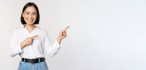 Portrait of beautiful asian woman, saleswoman pointing fingers right and showing info, banner or logo, sale advertisement, standing over white background - Photo, Image