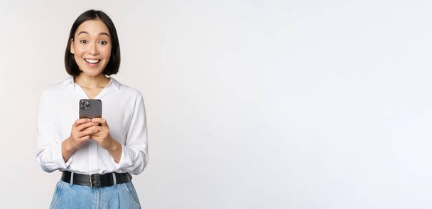 Excited asian woman smiling, reacting to info on mobile phone, holding smartphone and looking happy at camera, standing over white background - Photo, Image