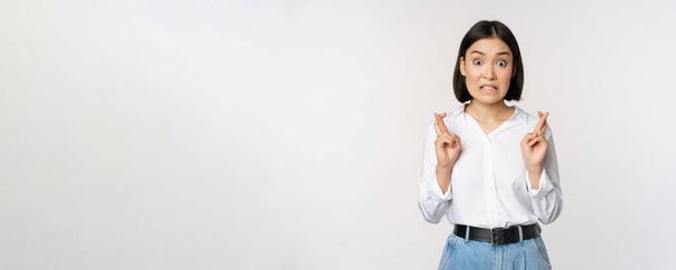 Retrato de mujer joven coreana, chica asiática cruzan los dedos y rezan, pidiendo deseo, anticipando, esperando resultados, de pie sobre fondo blanco - Foto, imagen