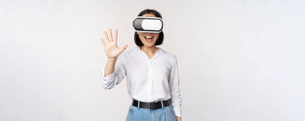 Mosolygó ázsiai nő integetett neked, visel virtuális valóság szemüveg, vr headset, csevegés a meta univerzumban, álló felett fehér háttér - Fotó, kép