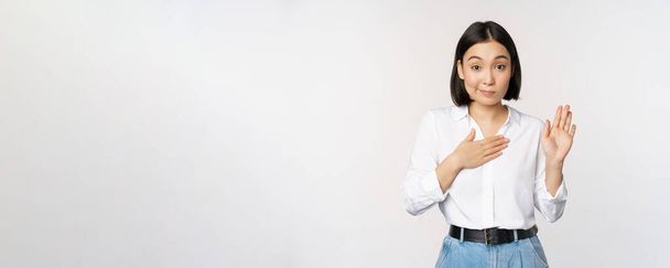 Imagen de la joven y linda trabajadora de oficina, estudiante asiática levantando la mano y poniendo la palma en el pecho, nombre de sí misma, introducir, haciendo promesa, de pie sobre fondo blanco - Foto, imagen
