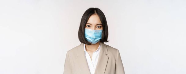 Coronavirus and workplace concept. Portrait of asian office worker, businesswoman in medical face mask looking at camera, white background - 写真・画像
