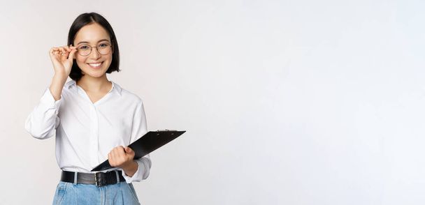 Jovem mulher, gerente de escritório trabalhador em óculos, segurando prancheta e olhando como profissional, de pé contra fundo branco - Foto, Imagem