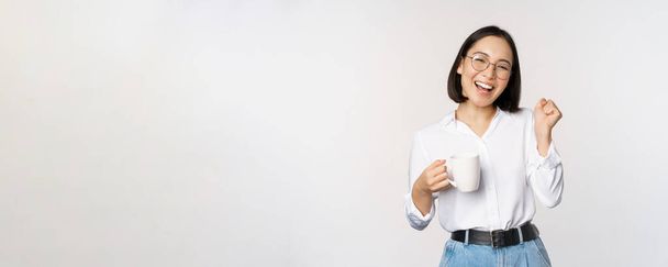 Happy dancing woman drinking coffee or tea from mug. Korean girl with cup, standing over white background - 写真・画像